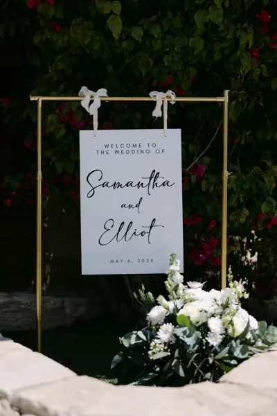 White welcome sign suspended in a black frame with white flowers and greenery at its base.
