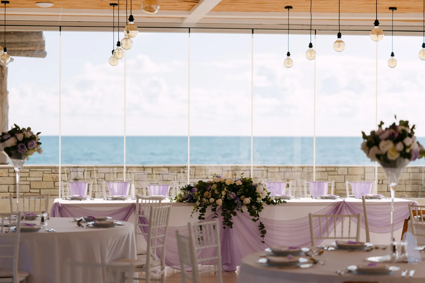 Sea-view reception with softly coloured lilac table linen, white chairs and raised floral centrepieces.