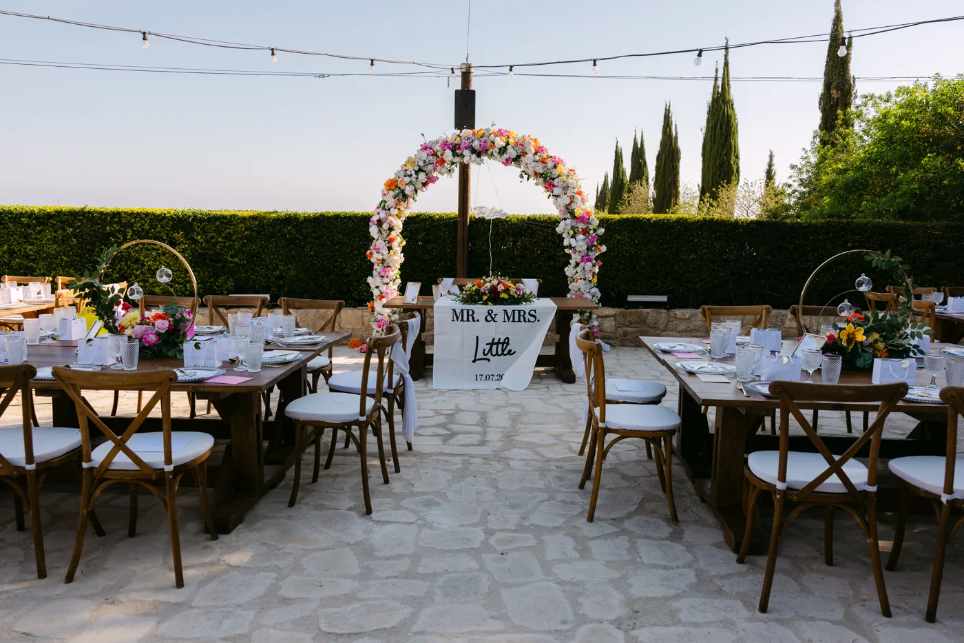 Wide reception terrace with wooden banquet tables and a colourful floral arch behind the sweetheart table.