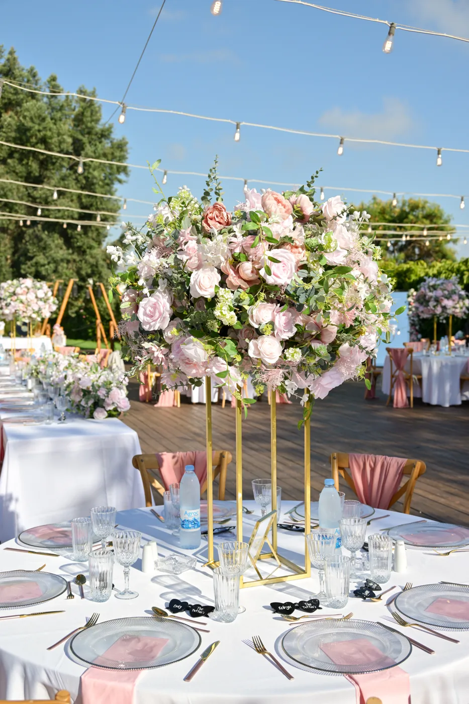 Large mixed pastel floral arrangement on a slender gold stand above a set outdoor reception table.