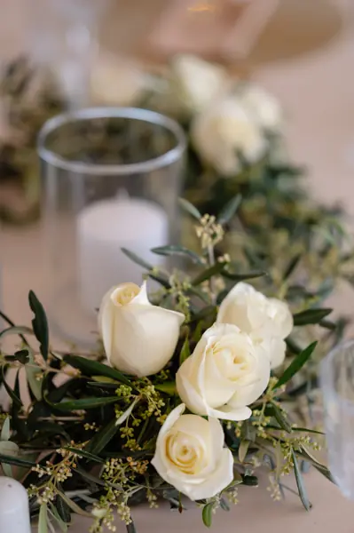White candle in a glass cylinder surrounded by white roses and green foliage.